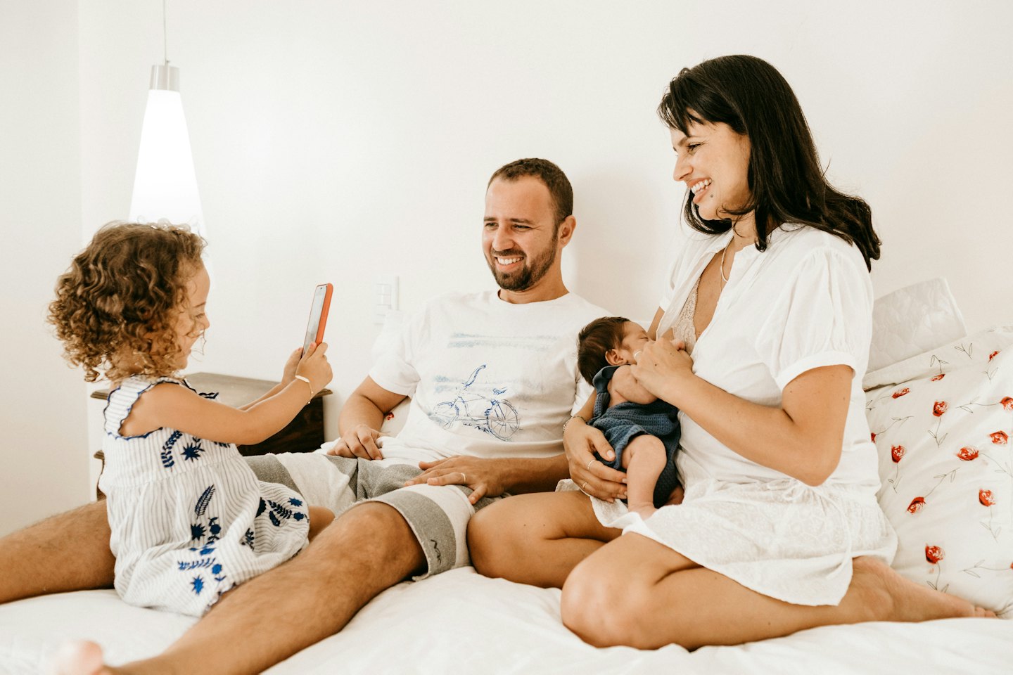 Smiling woman breastfeeding a baby while man stands nearby and toddler holds phone.